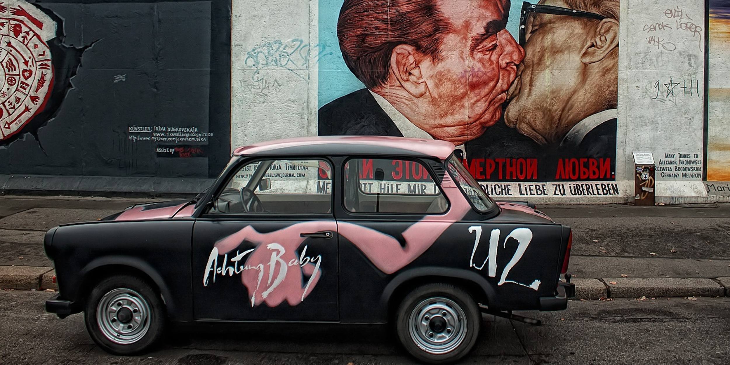 Berlin with an artwork in the background and an old black car with the words Achtung Baby painted on it